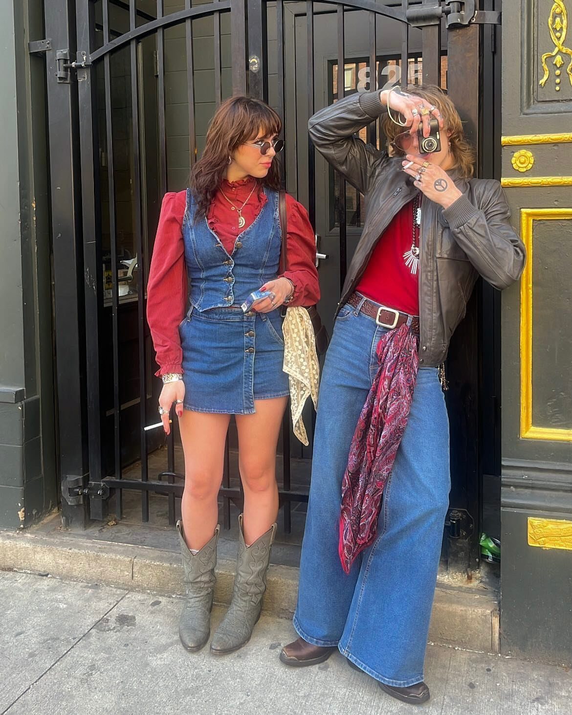 70s Inspired Outfits with 22 Groovy Looks to Try Now 21 Two women posing in retro-style denim outfits and red tops in front of a black gate.