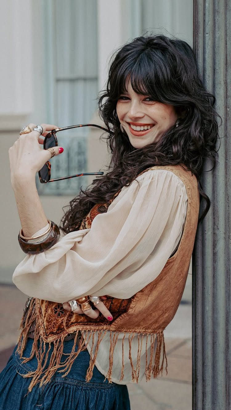 70s Inspired Outfits with 22 Groovy Looks to Try Now 3 Smiling woman with dark wavy hair wearing a cream blouse and a brown fringed suede vest.