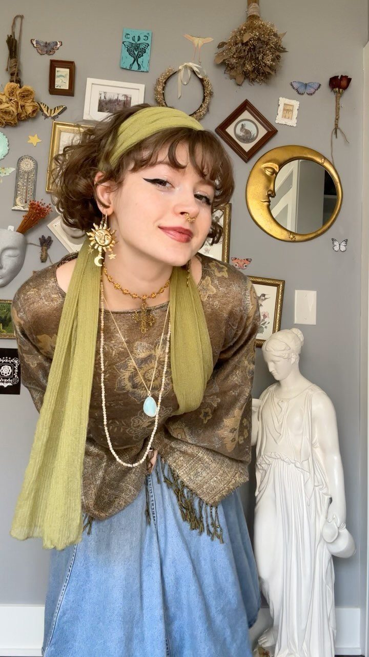 70s Inspired Outfits with 22 Groovy Looks to Try Now 22 A young woman posing in a gold patterned blouse, green headband, and layered jewelry against a decorated wall.
