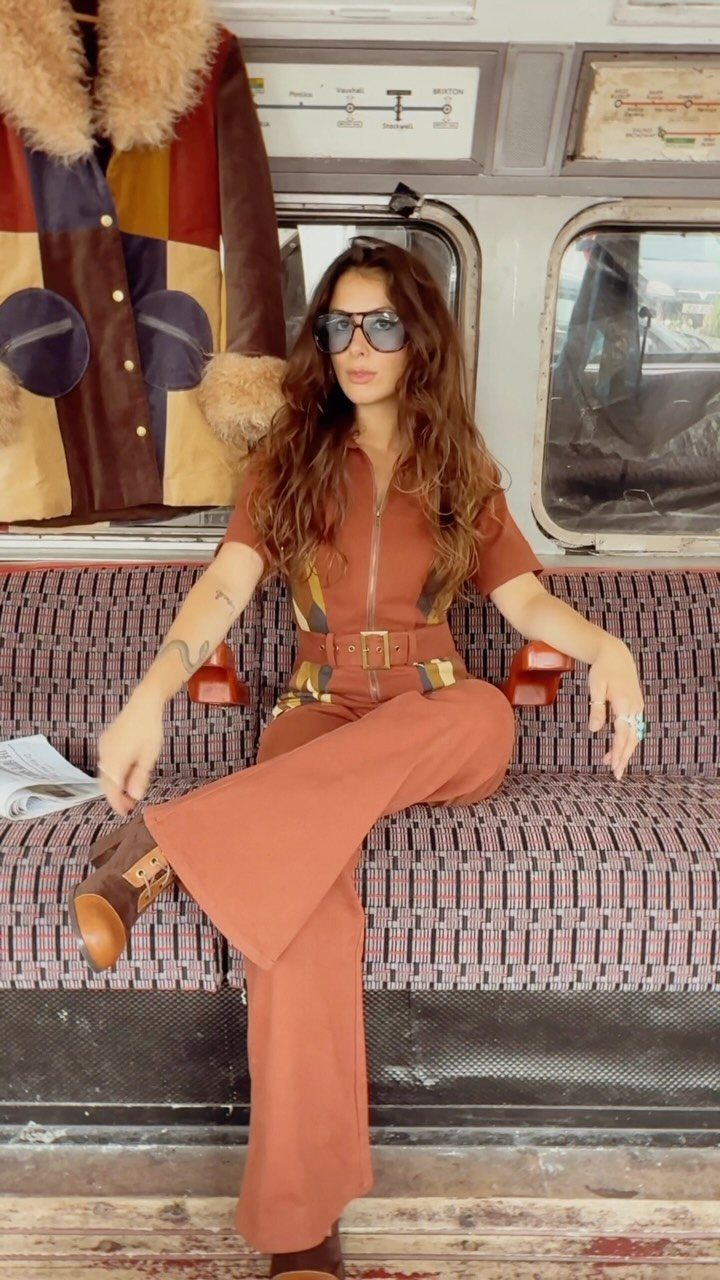 70s Inspired Outfits with 22 Groovy Looks to Try Now 2 Woman in a burnt orange jumpsuit and sunglasses sitting on a patterned bench inside a vintage van.
