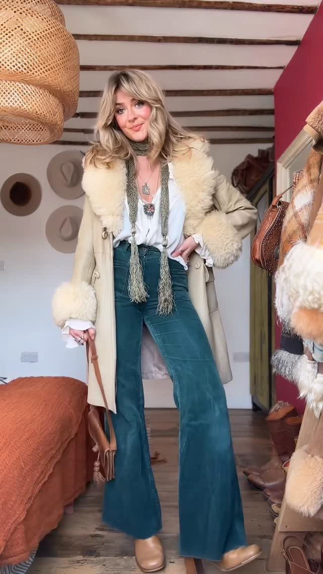 70s Inspired Outfits with 22 Groovy Looks to Try Now 10 Woman wearing a shearling coat, white blouse, and teal flared jeans standing indoors.