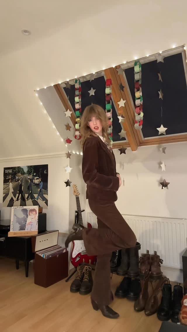 70s Inspired Outfits with 22 Groovy Looks to Try Now 14 Woman posing in a brown velvet flared outfit with a guitar and records in the background.