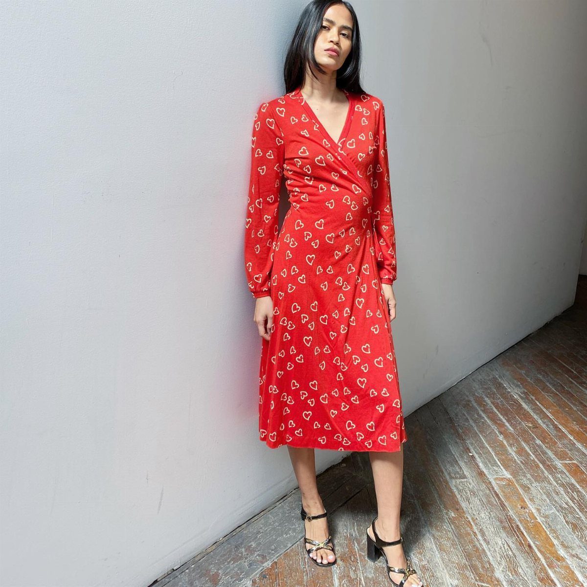 70s Inspired Outfits with 22 Groovy Looks to Try Now 1 Woman in a red long-sleeved wrap dress with white heart patterns standing against a wall.