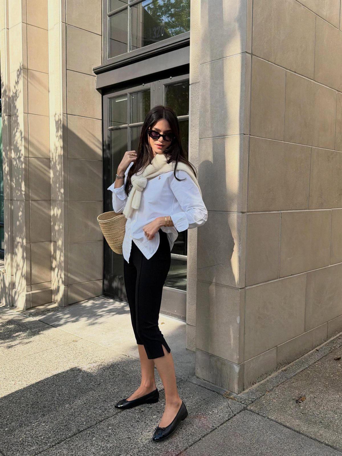 Woman wearing white button-down shirt, black cropped trousers, and a sweater over her shoulders.