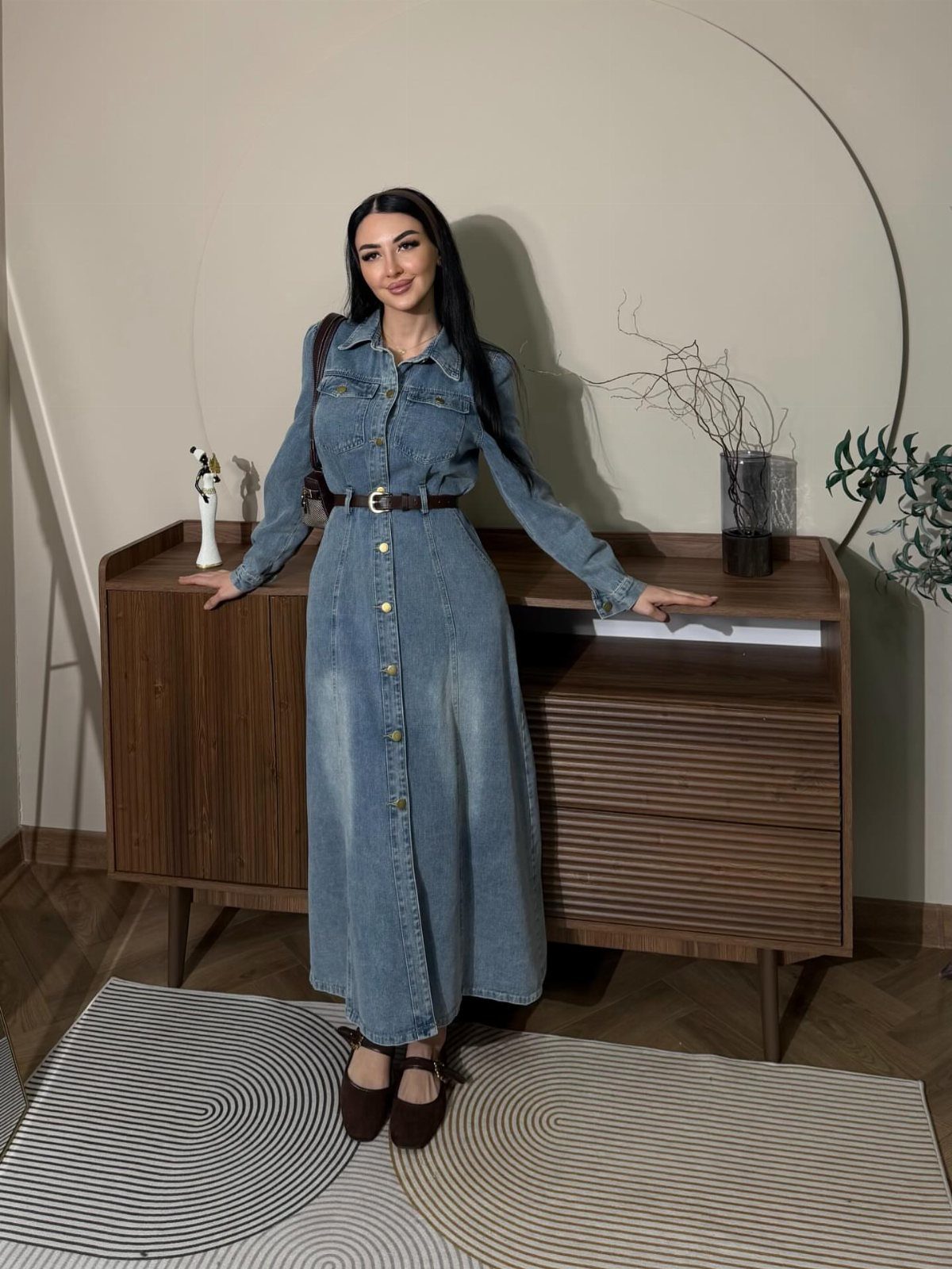 Woman wearing a long blue denim shirtwaist dress with a brown belt and shoes.