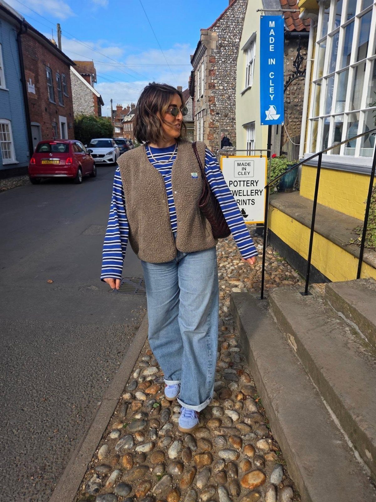 Woman wearing a brown knit vest, striped shirt, and light-wash jeans on a street.