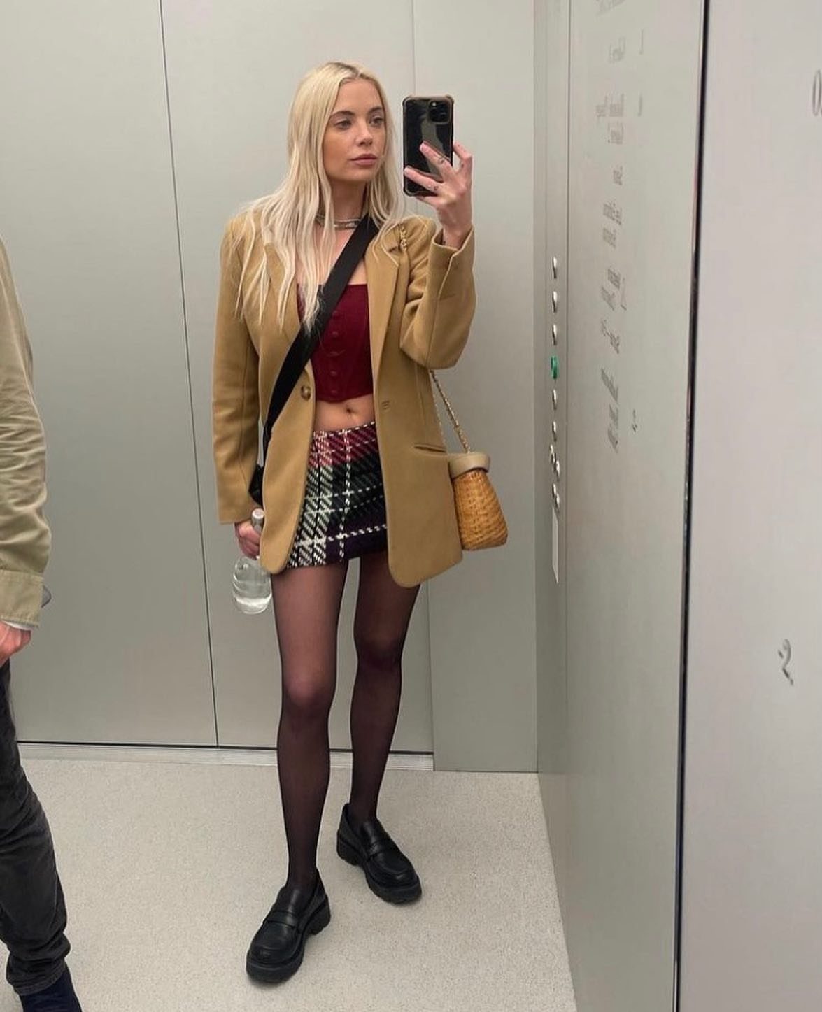 Woman in tan blazer and plaid mini skirt taking a mirror selfie in elevator