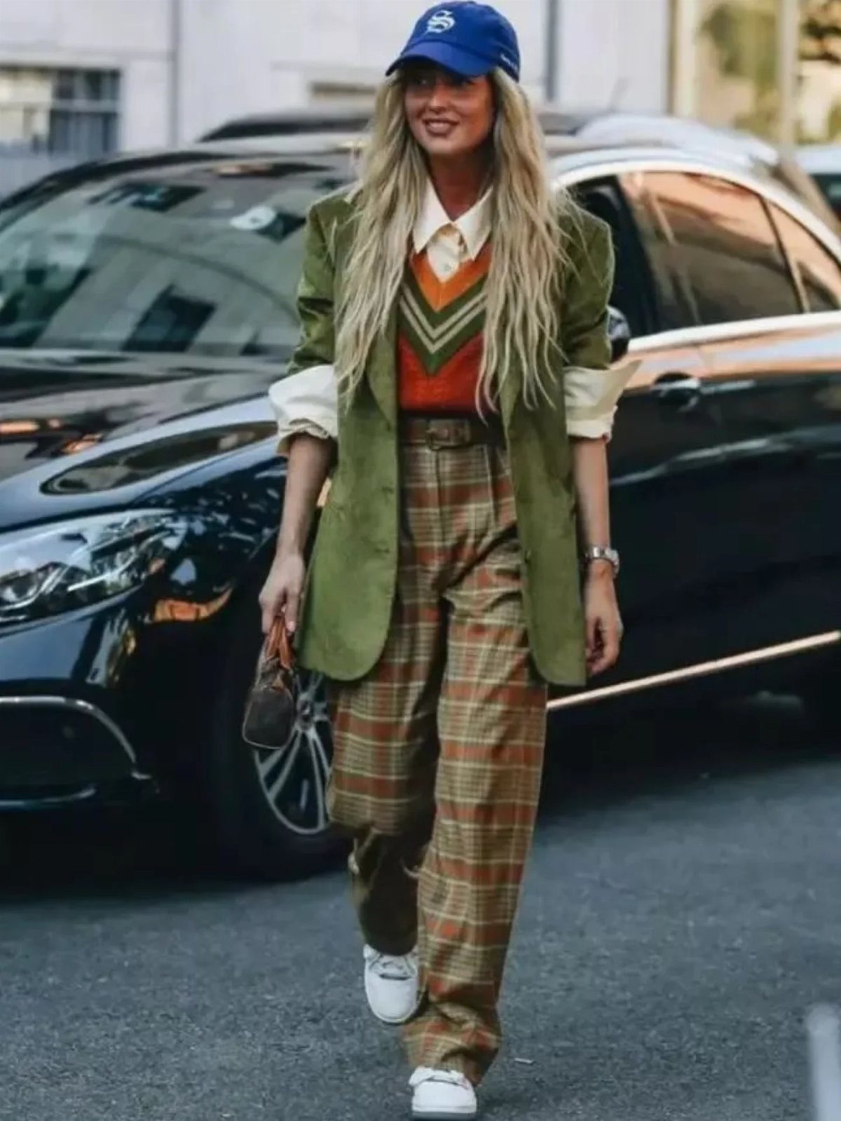 Woman in green blazer and brown plaid trousers posing by a black car.