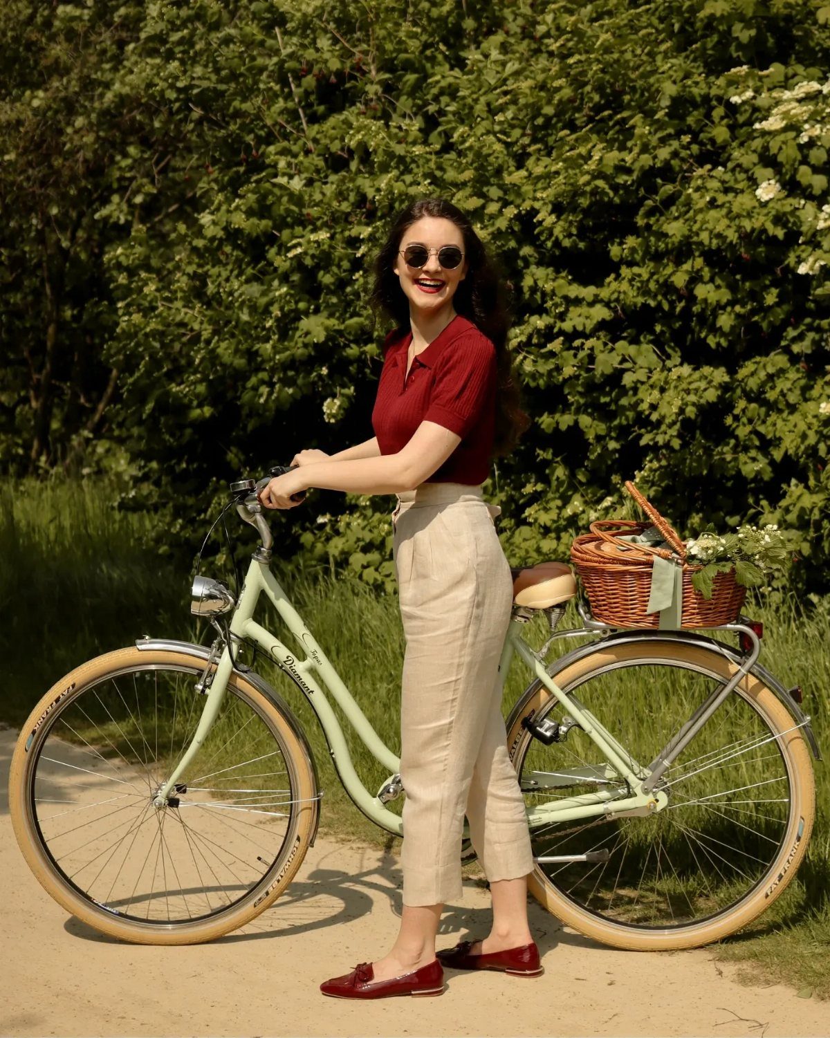 Woman in red knit polo and beige pleated trousers posing with a green bicycle.