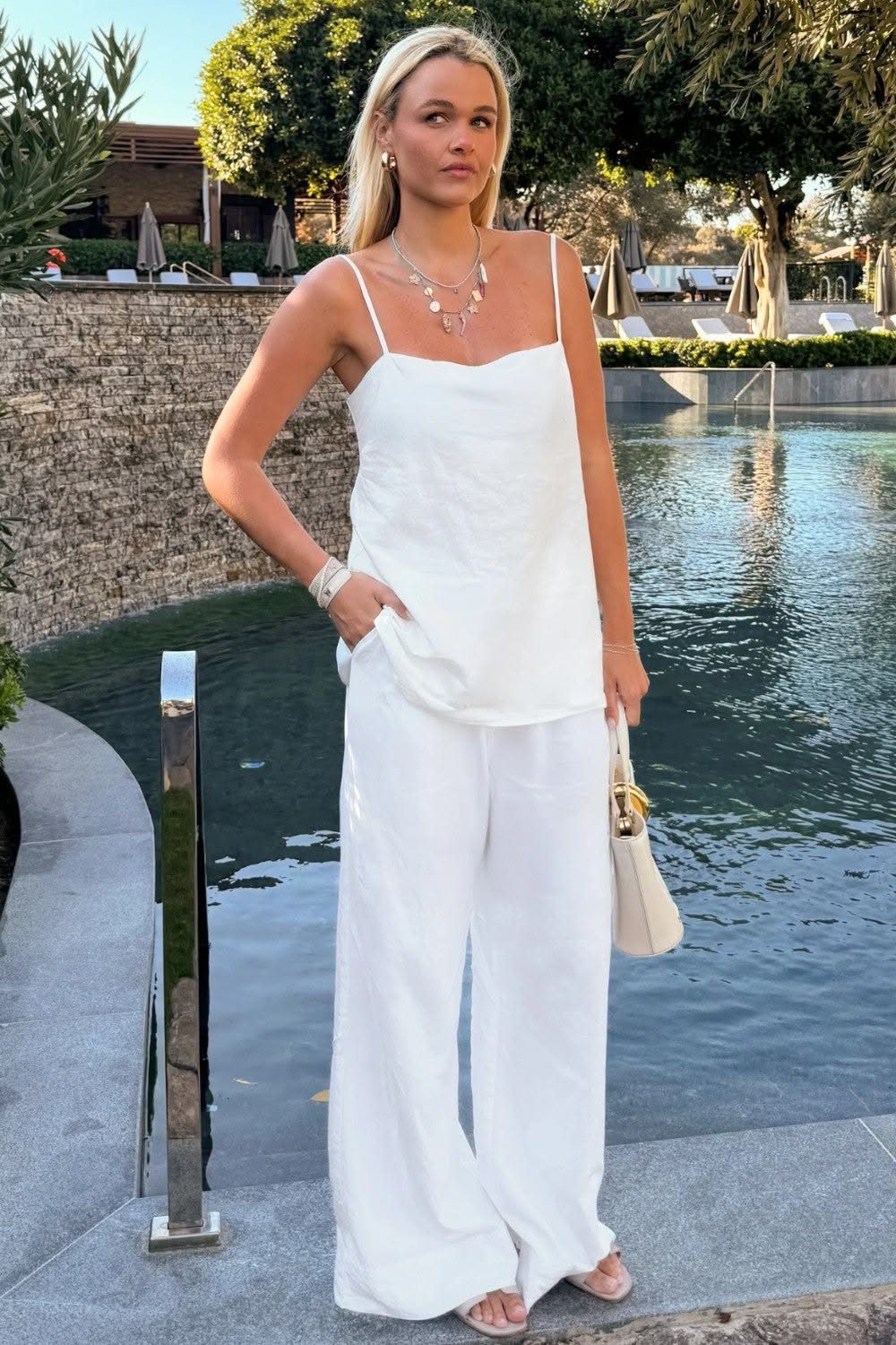 Blonde woman posing in a white camisole and wide-leg white linen trousers by a swimming pool.