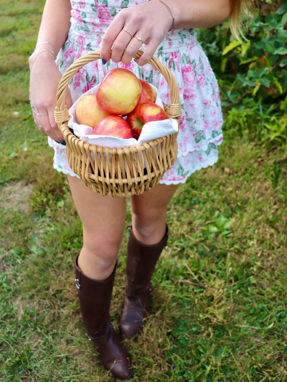 Apple Picking Outfit Ideas: 12 Looks for the Orchard 8 Woman in floral dress and brown boots holding a basket of red apples.