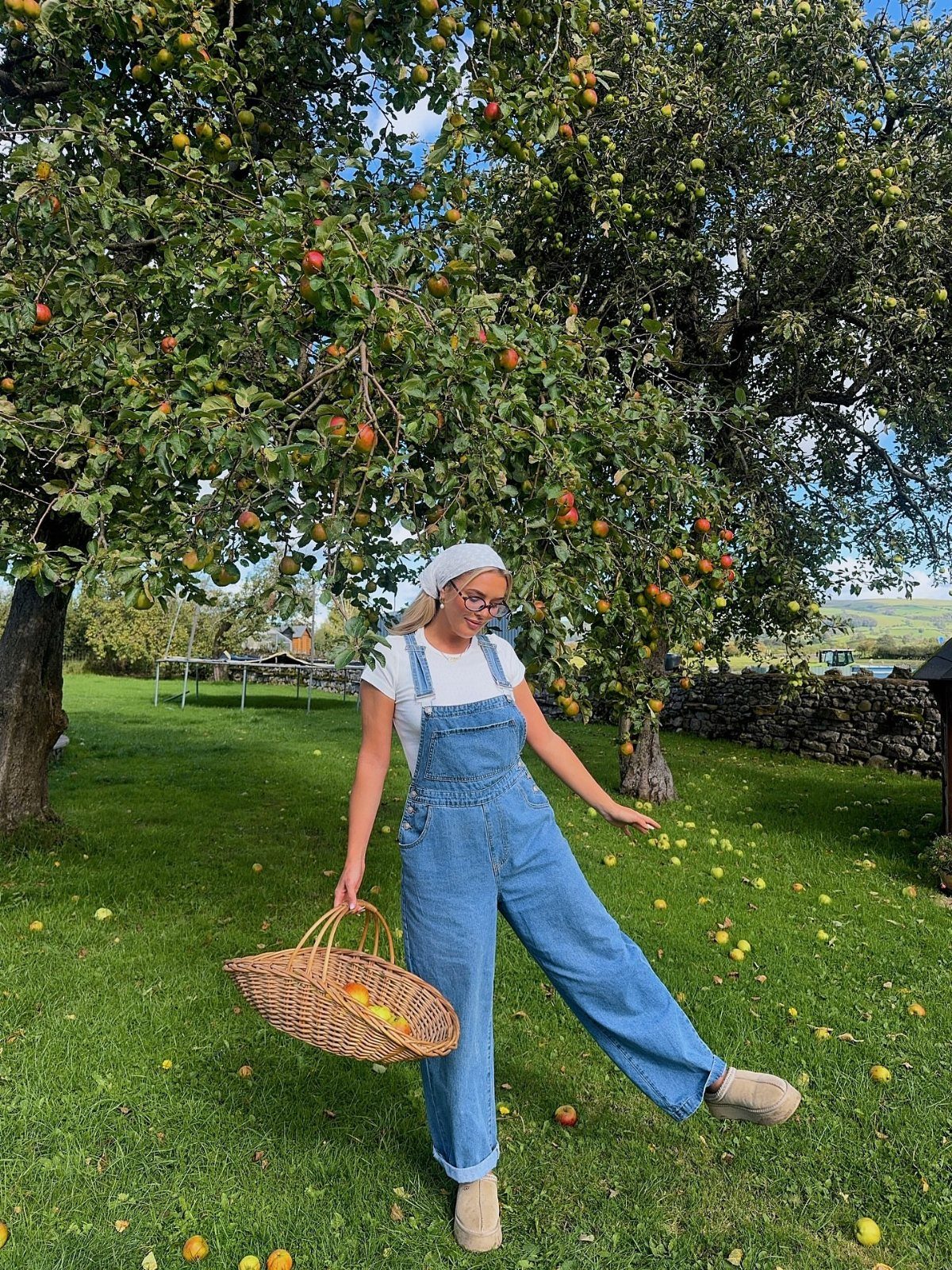 Apple Picking Outfit Ideas: 12 Looks for the Orchard 4 Woman in denim overalls and beige mules holding a basket of apples in an orchard.