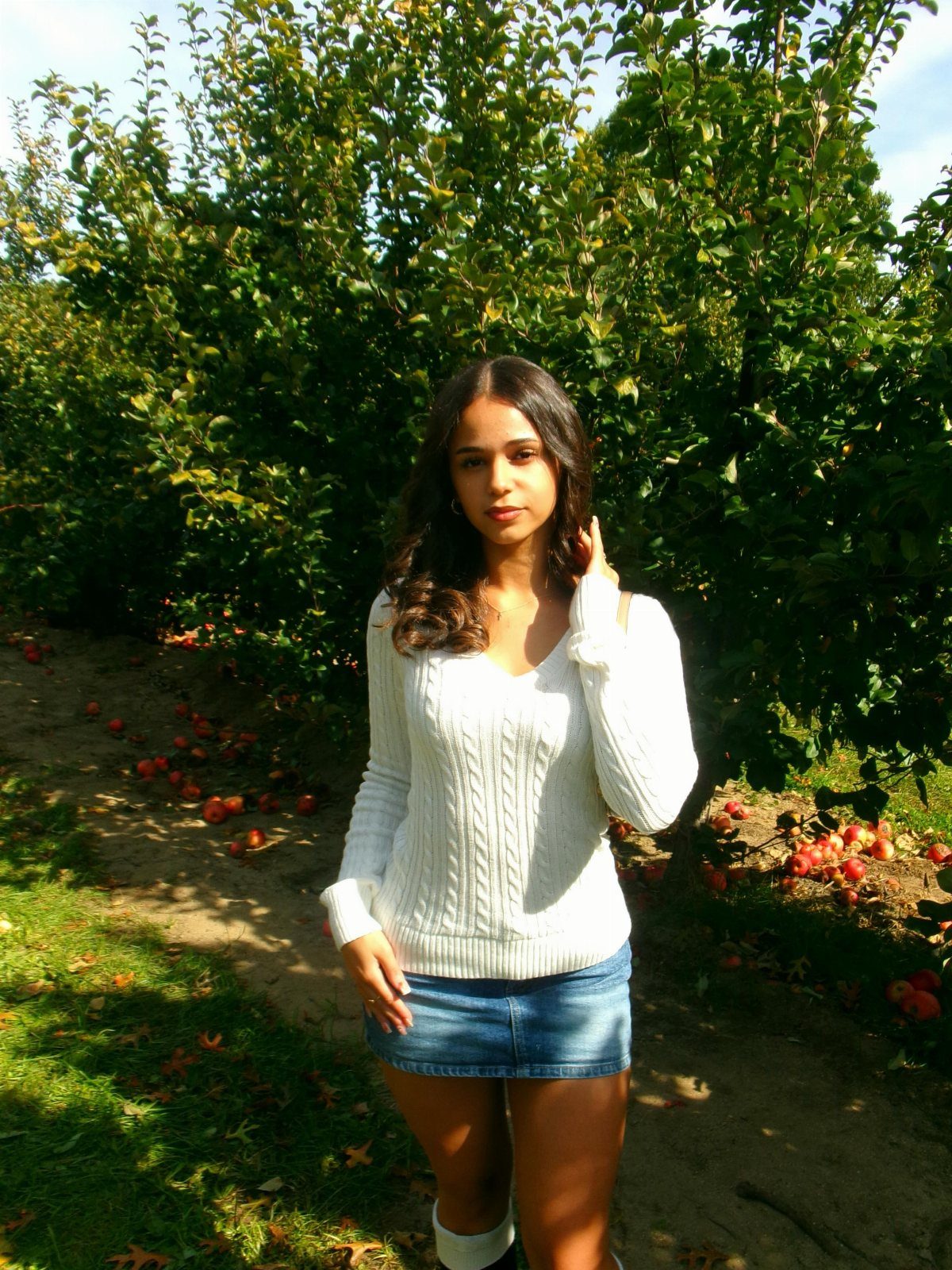 Apple Picking Outfit Ideas: 12 Looks for the Orchard 10 Woman in white cable-knit sweater and denim skirt posing in an apple orchard.