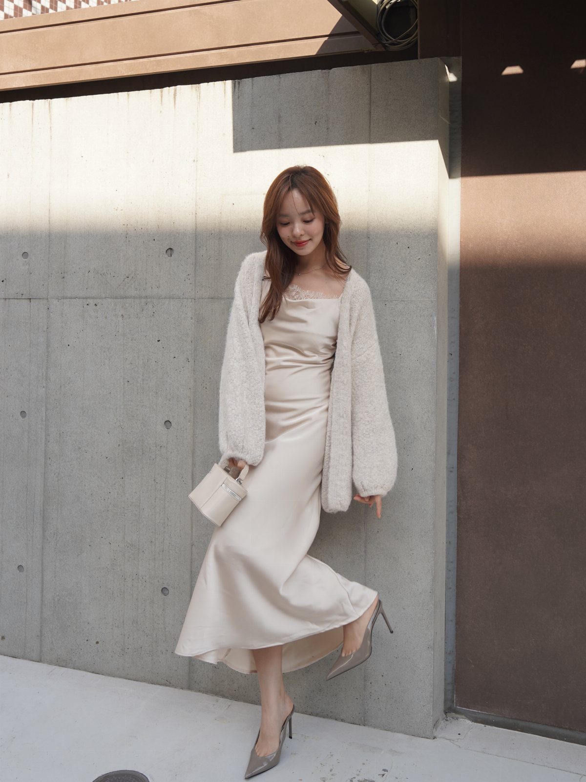 Woman in a beige oversized cardigan and white satin slip dress posing by a wall.