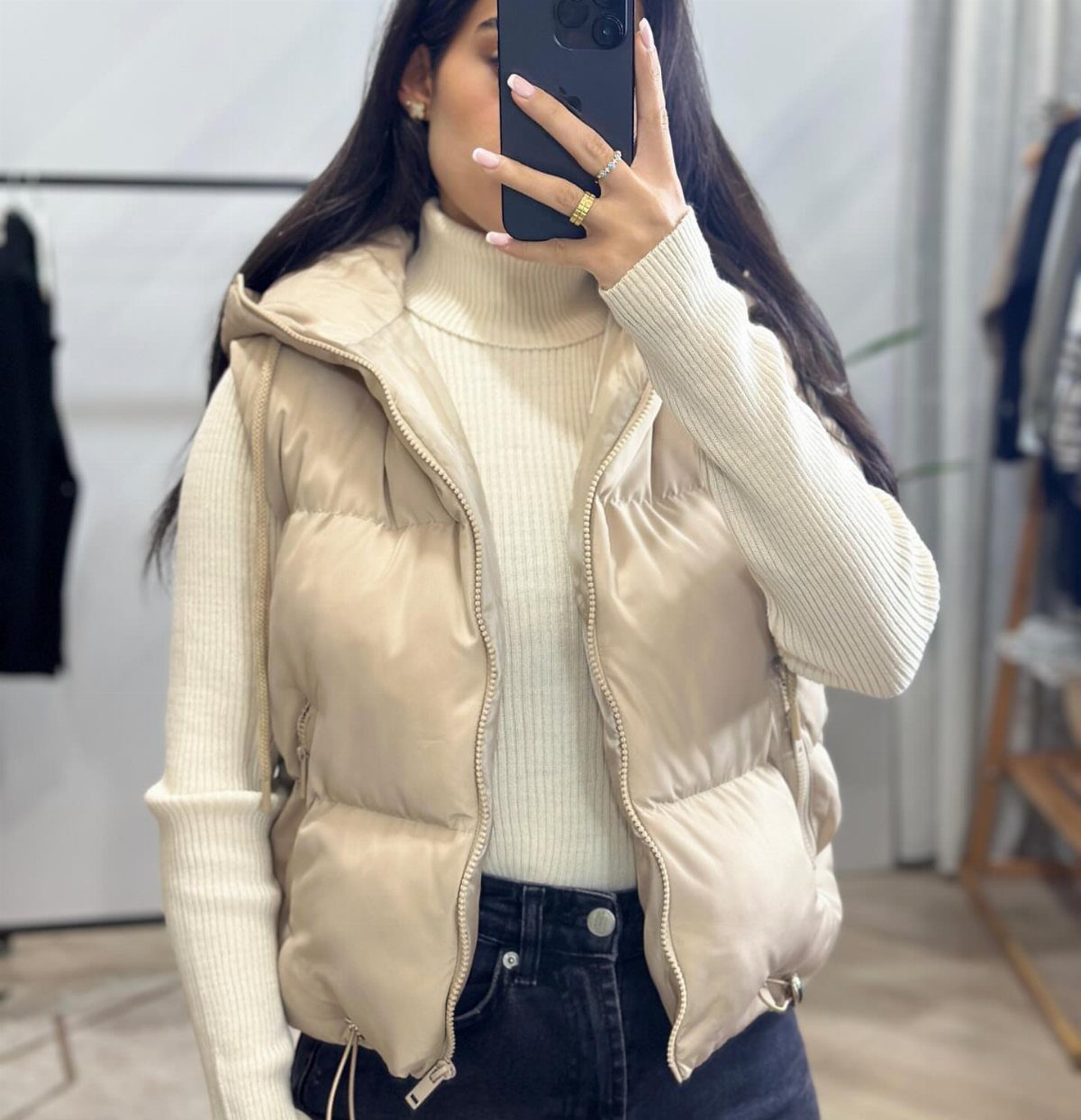Woman wearing a beige puffer vest and white ribbed turtleneck sweater with dark jeans.