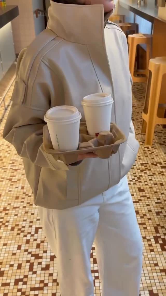Beige leather jacket and white trousers worn by a person holding coffee cups.