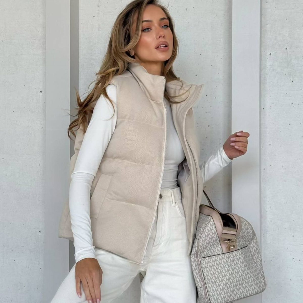 Woman wearing a beige puffer vest and white jeans with a patterned handbag.