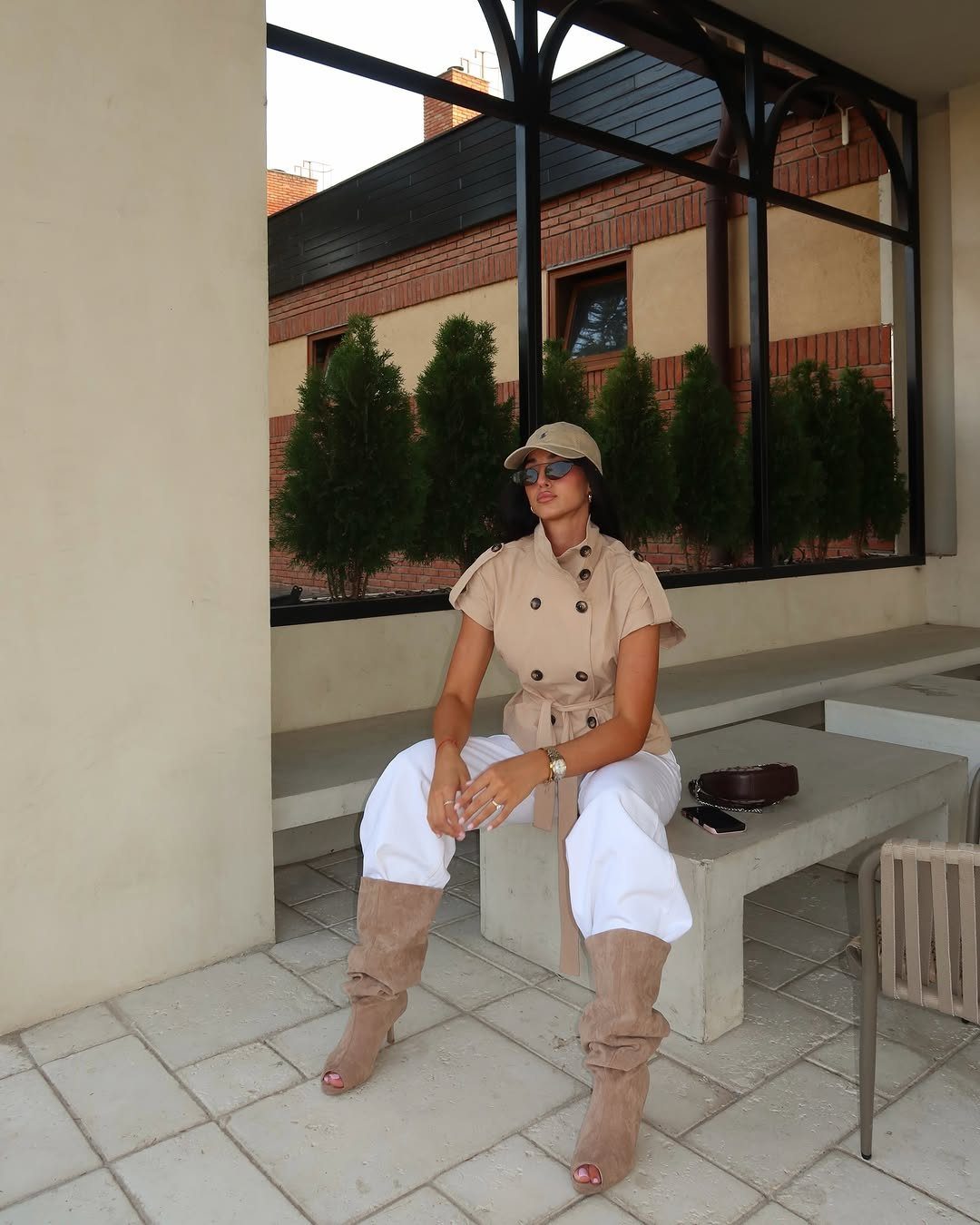 Woman in a beige belted vest and white trousers sitting on a concrete bench.