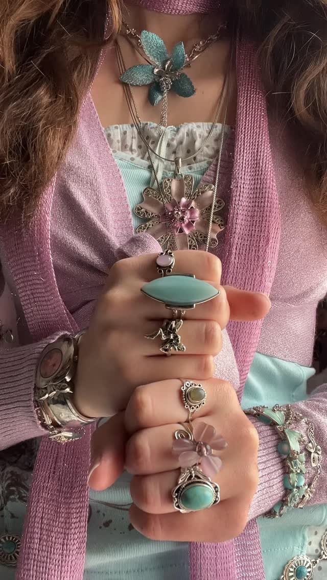 Close up of hands wearing large turquoise statement rings and layered necklaces with floral pendants.