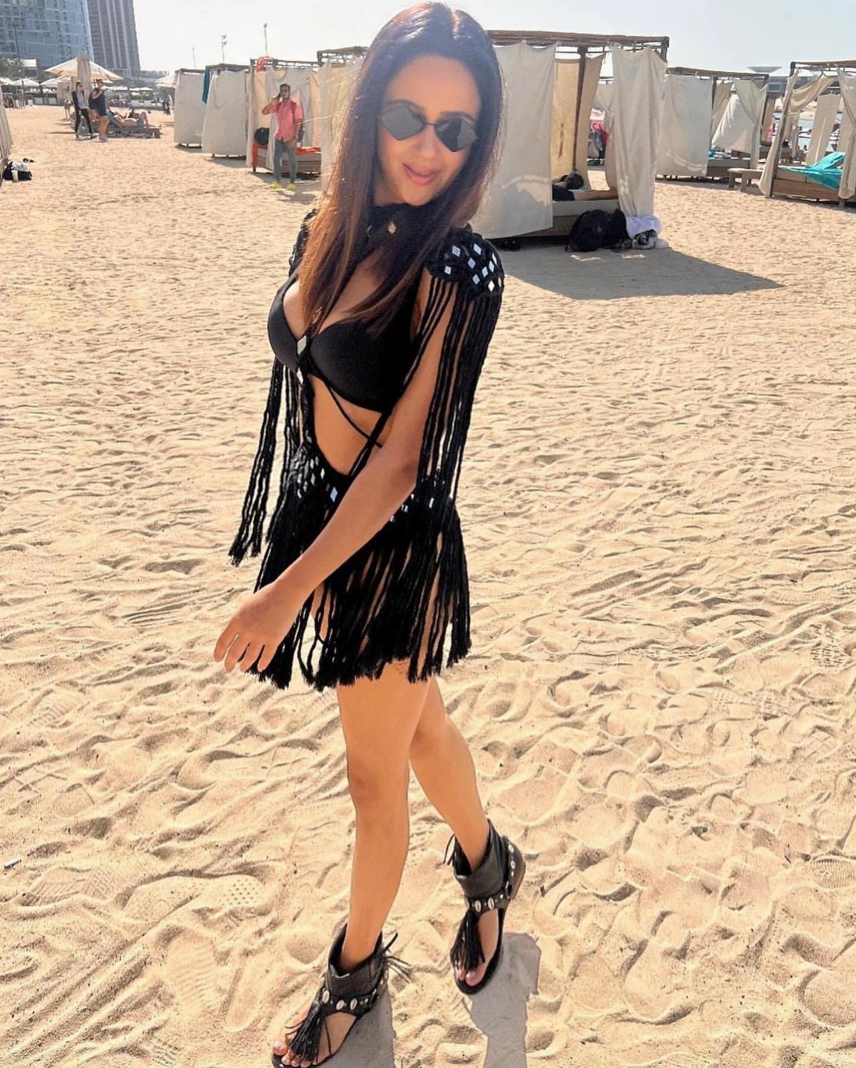 Woman in a black fringed beach cover-up walking on a sunny sandy beach.