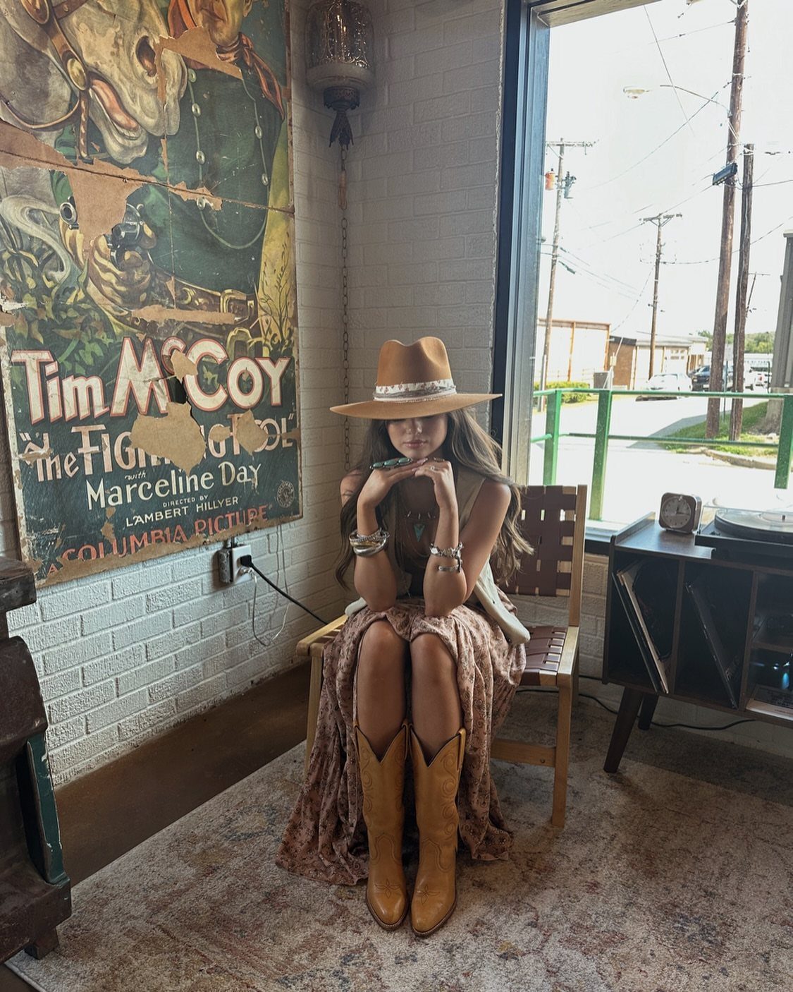 Woman in boho attire wearing a wide-brimmed hat and tan boots sitting on a chair.
