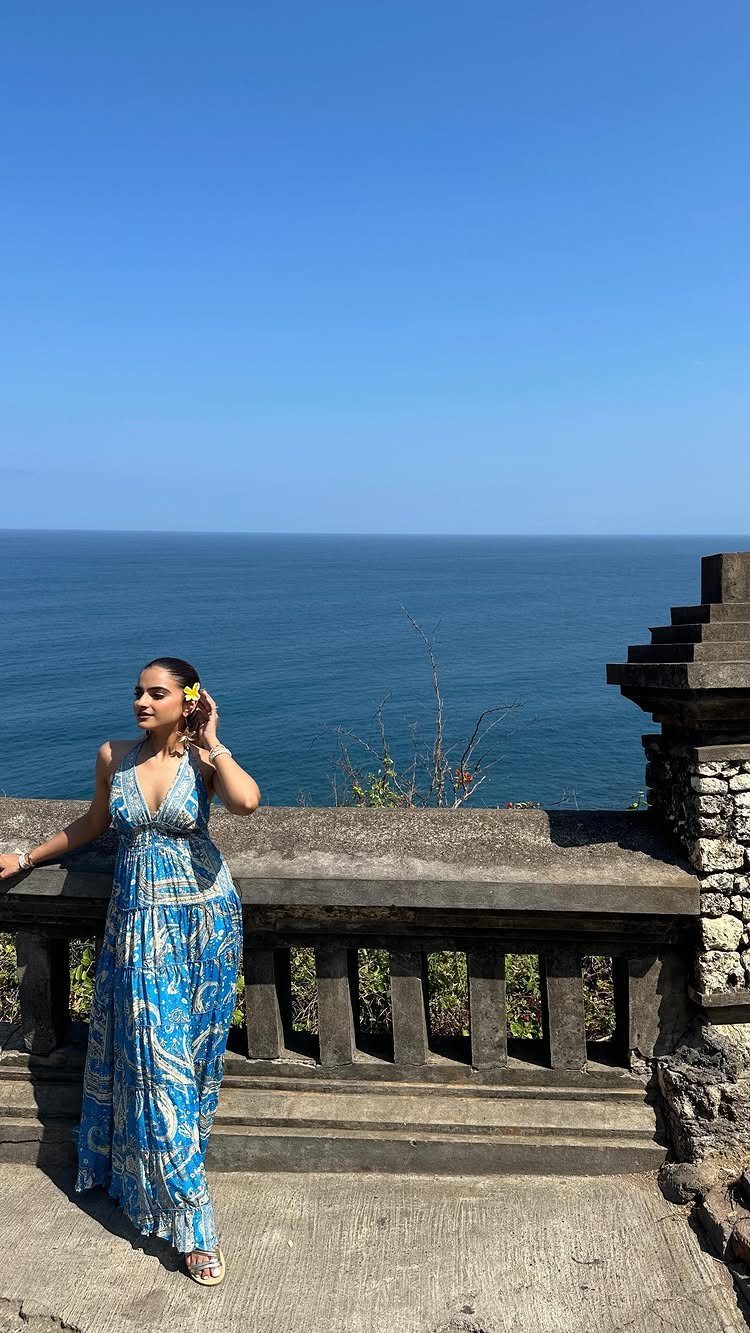 Woman wearing a blue patterned flowy maxi dress posing by a stone railing overlooking the ocean.