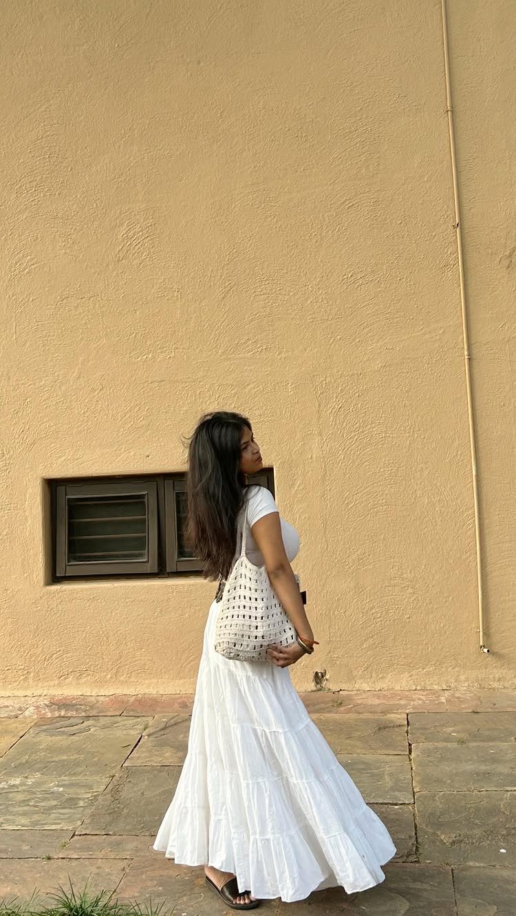 Woman in a white top and flowing white tiered maxi skirt standing by a yellow wall.