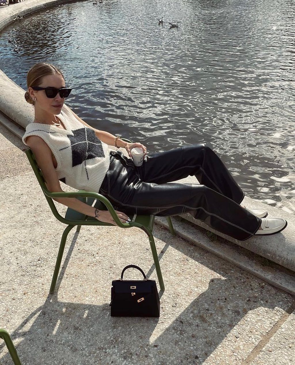 Woman in black leather trousers and knit vest sitting by a lake.