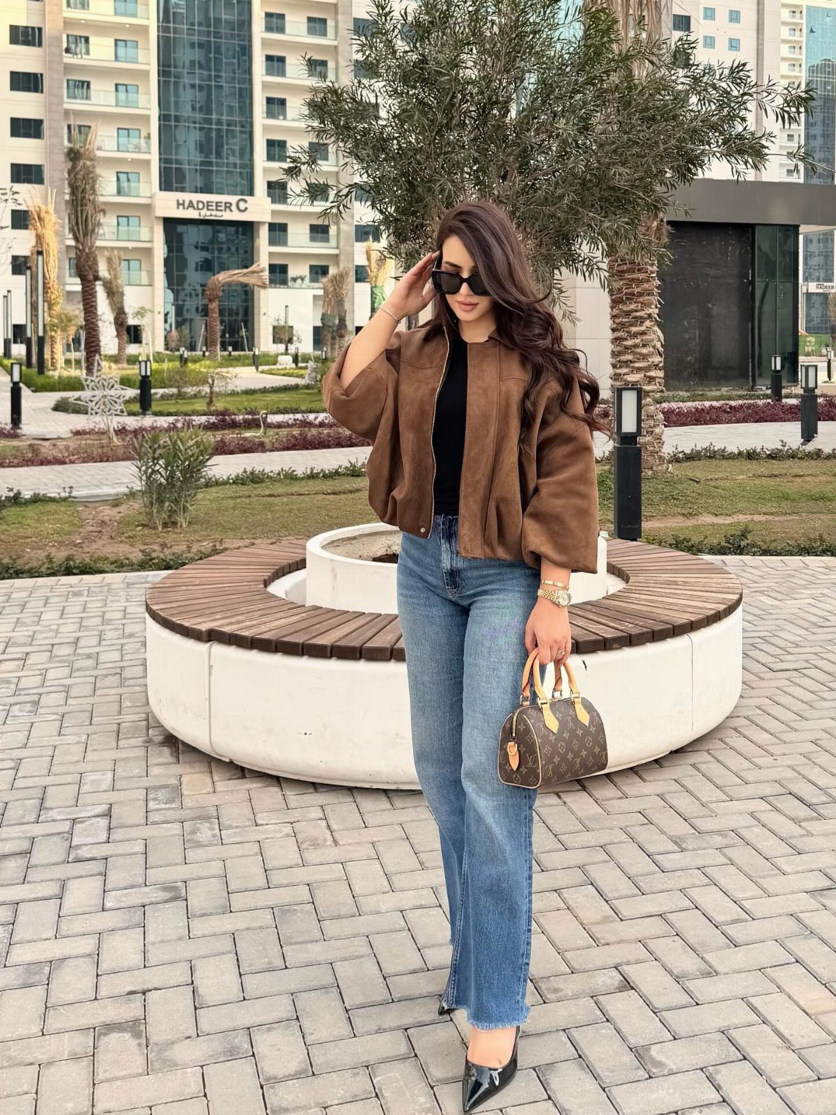 Woman wearing a brown light jacket, black top, and blue jeans in an urban setting.