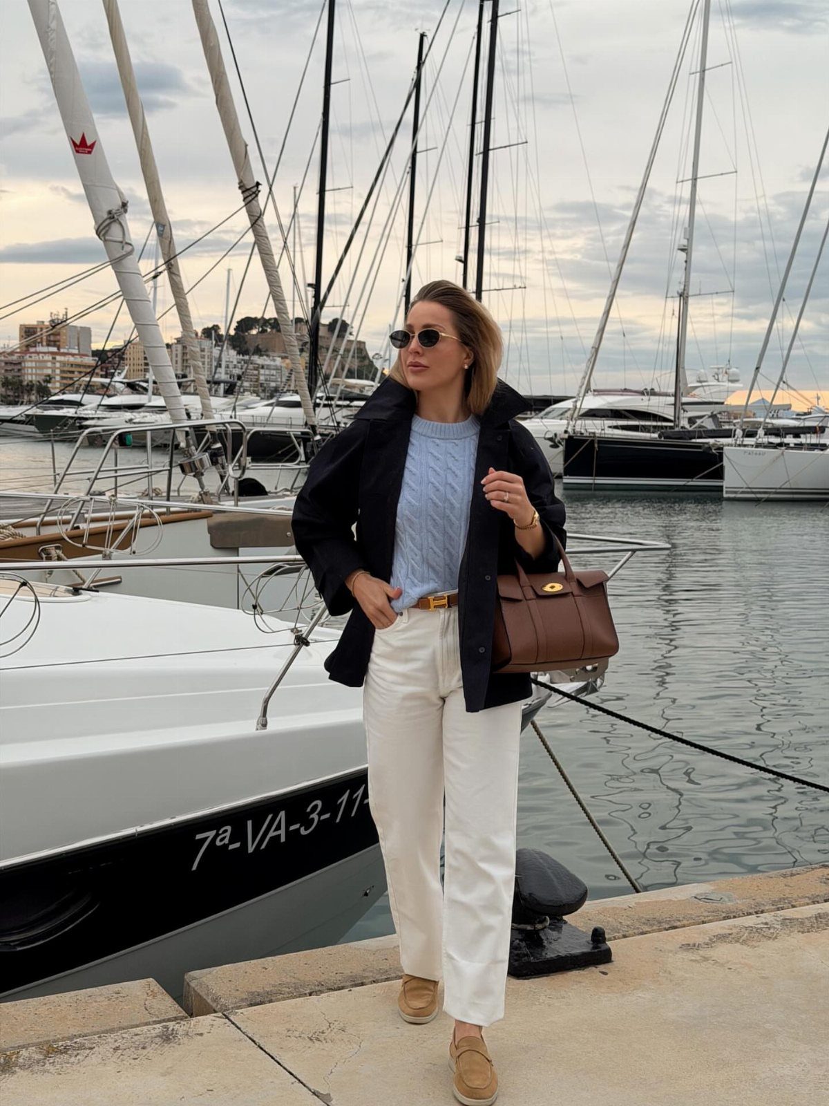 Woman wearing a black jacket, blue sweater, and white pants by a marina.