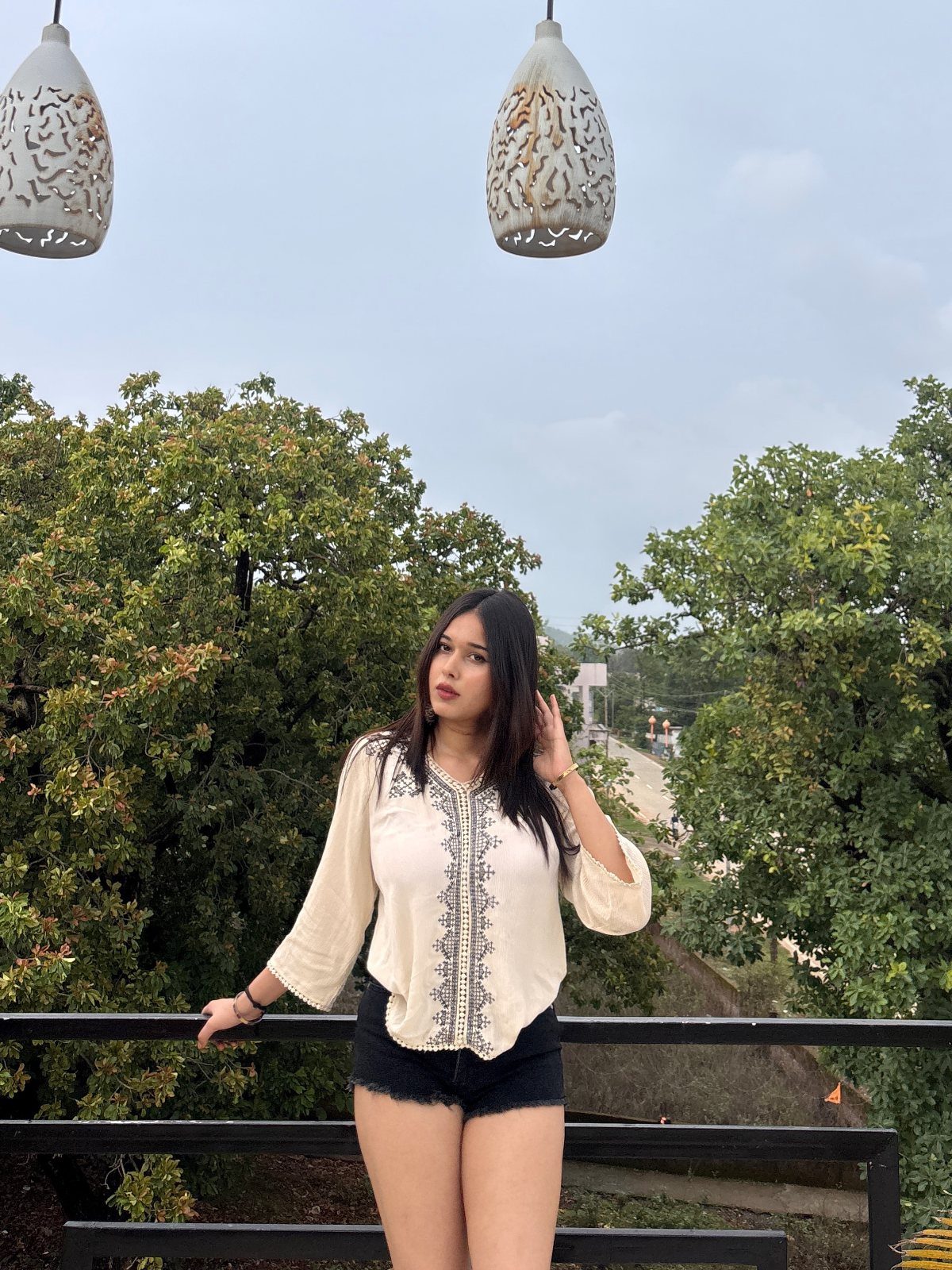 Woman wearing a cream embroidered blouse and black shorts standing outdoors near trees.