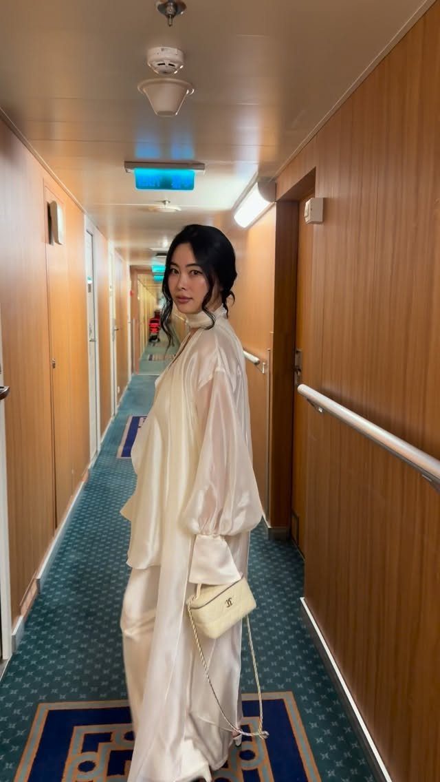 Woman in a long cream silky dress and white bag in a ship hallway.