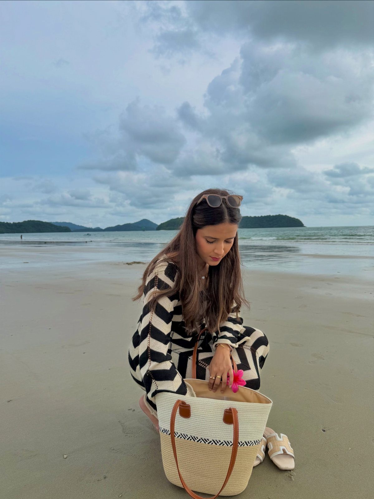 Woman in black and white striped outfit sitting on beach with straw tote bag