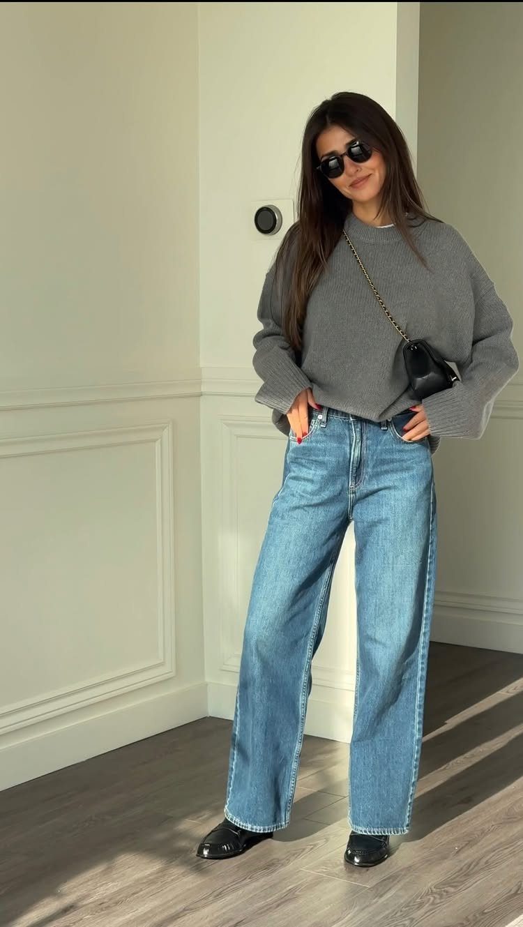 Woman wearing a grey sweater, wide-leg blue jeans, and black loafers.