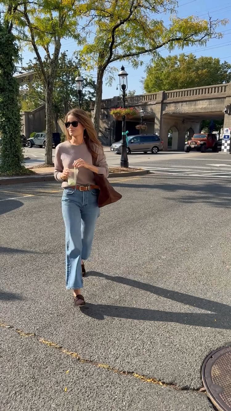 Woman wearing a brown sweater and light blue wide-leg jeans walking on a street.