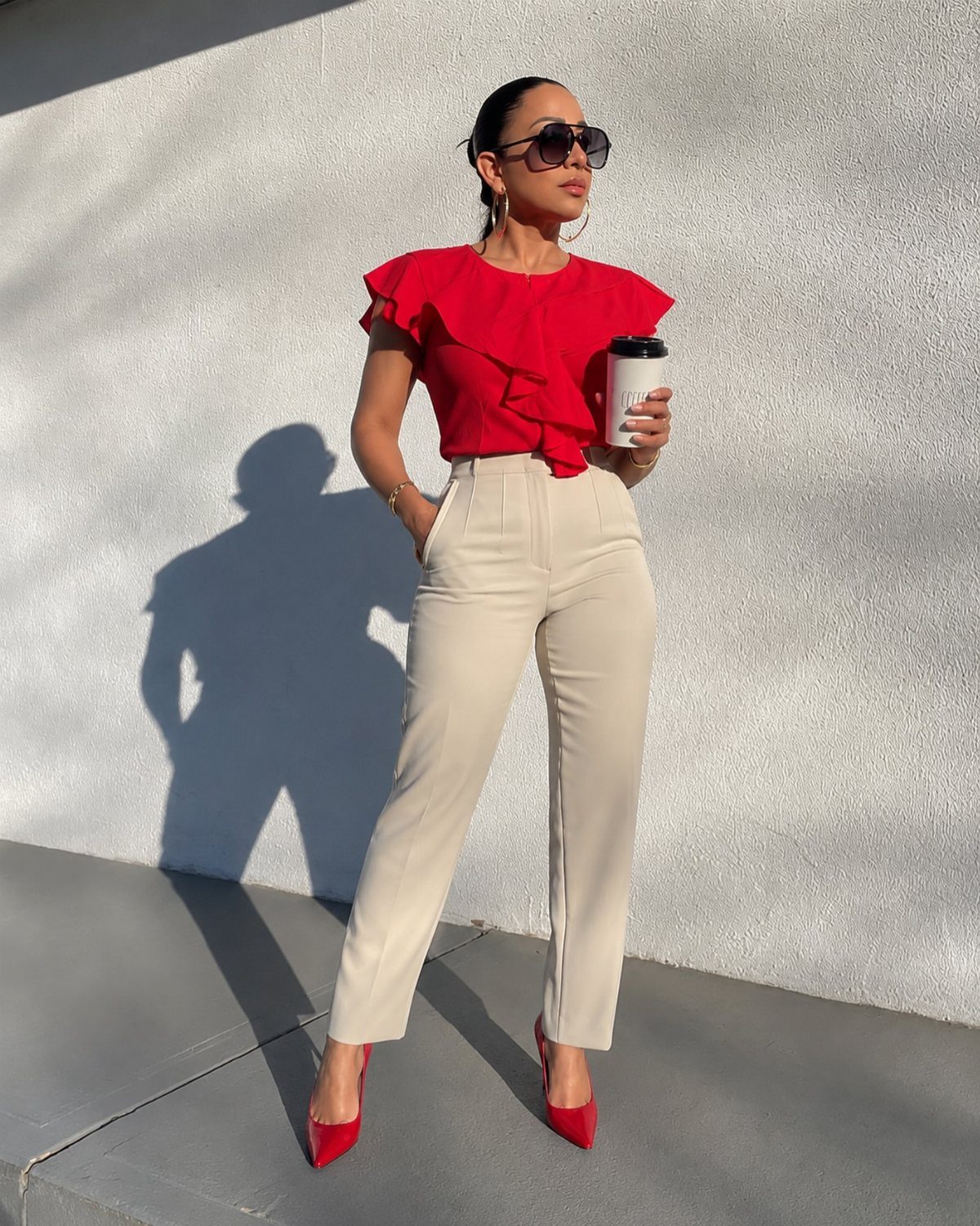 Woman in red ruffled blouse, beige trousers, and red heels holding a coffee cup.