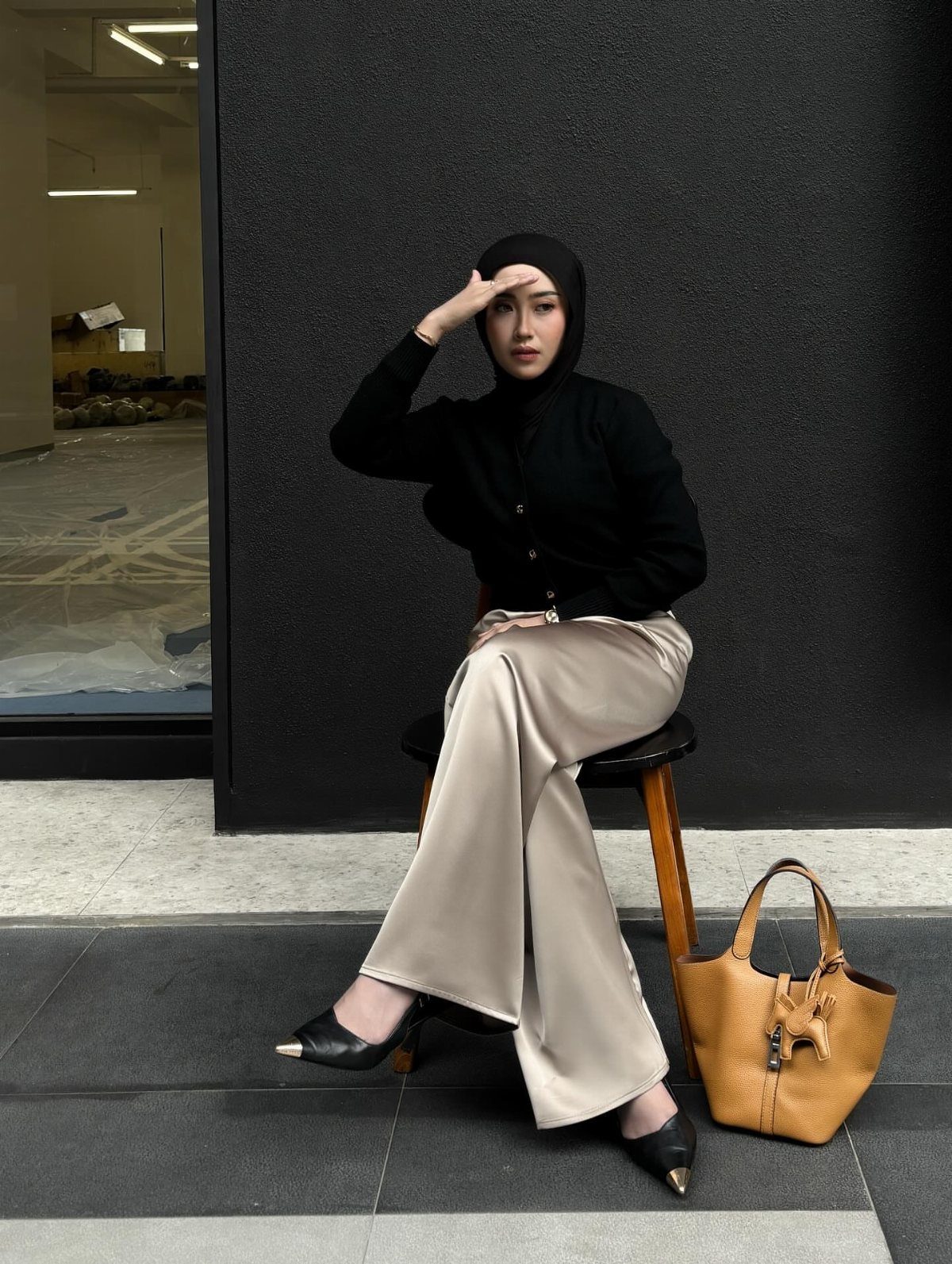 Woman in black cardigan and champagne satin wide-leg trousers sitting on a wooden stool.