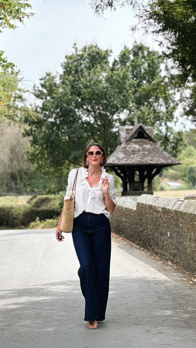 Woman wearing a white ruffled blouse and dark blue wide-leg jeans on a path.