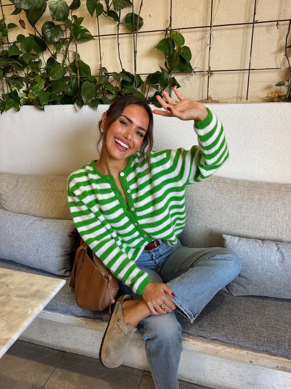 11 Casual Travel Outfit Ideas for Women (2026) 5 Woman wearing a green and white striped cardigan and blue jeans sitting on a sofa.