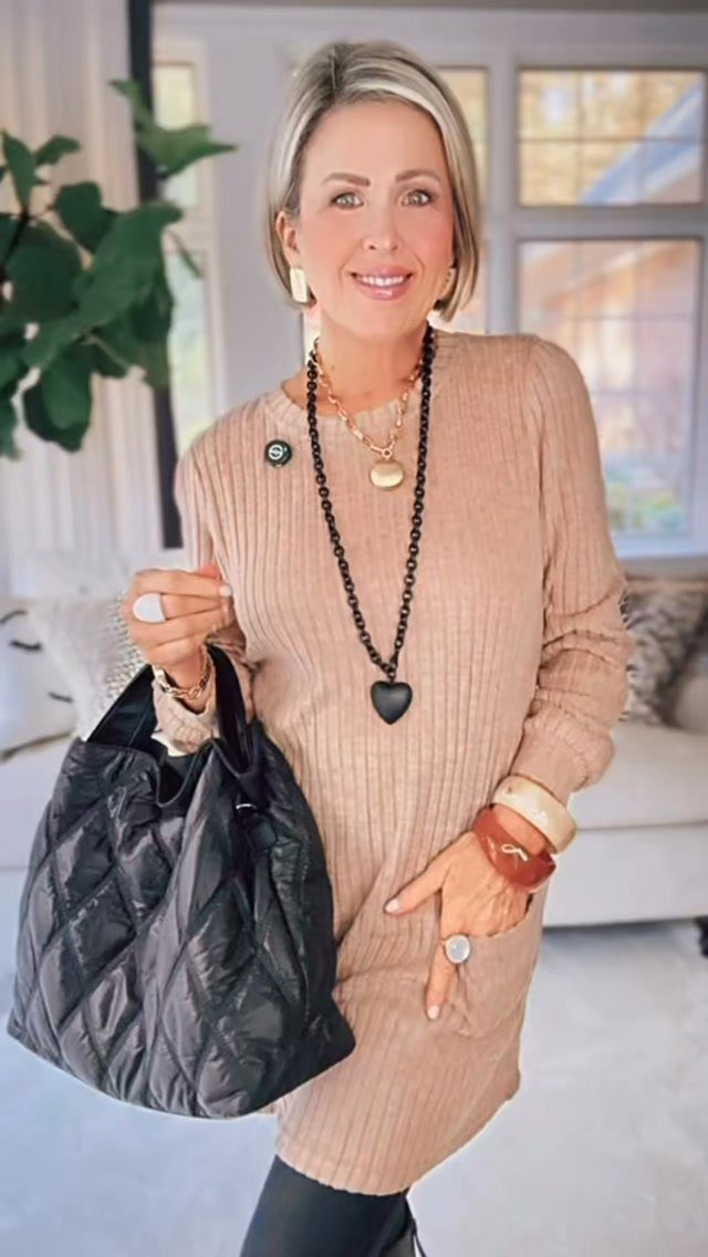 11 Casual Travel Outfit Ideas for Women (2026) 7 Woman wearing an oversized beige ribbed tunic sweater with black leggings and tote.