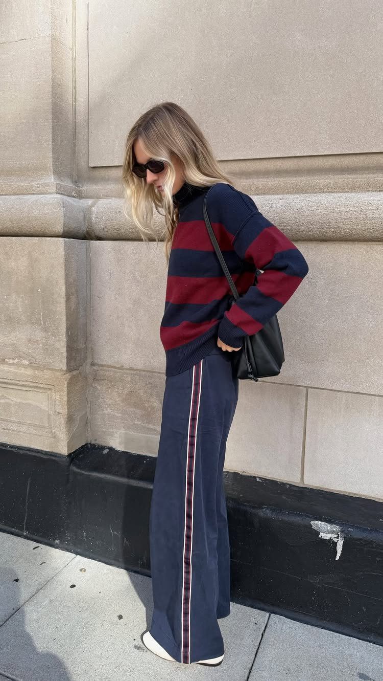 11 Casual Travel Outfit Ideas for Women (2026) 3 Woman in striped sweater, navy track pants, and white flats standing by a wall.