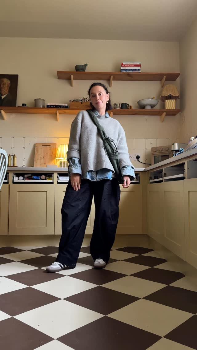 11 Casual Travel Outfit Ideas for Women (2026) 2 Woman wearing a grey sweater, blue shirt, and black wide-leg pants in a kitchen.