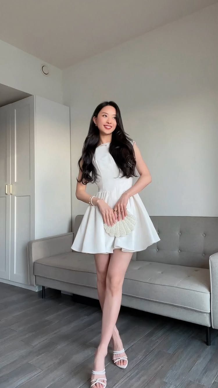 10 Chic Summer Outfits: Sandals, Linen, Dresses and More 3 A woman in a short white sleeveless dress posing in a room with a grey sofa.