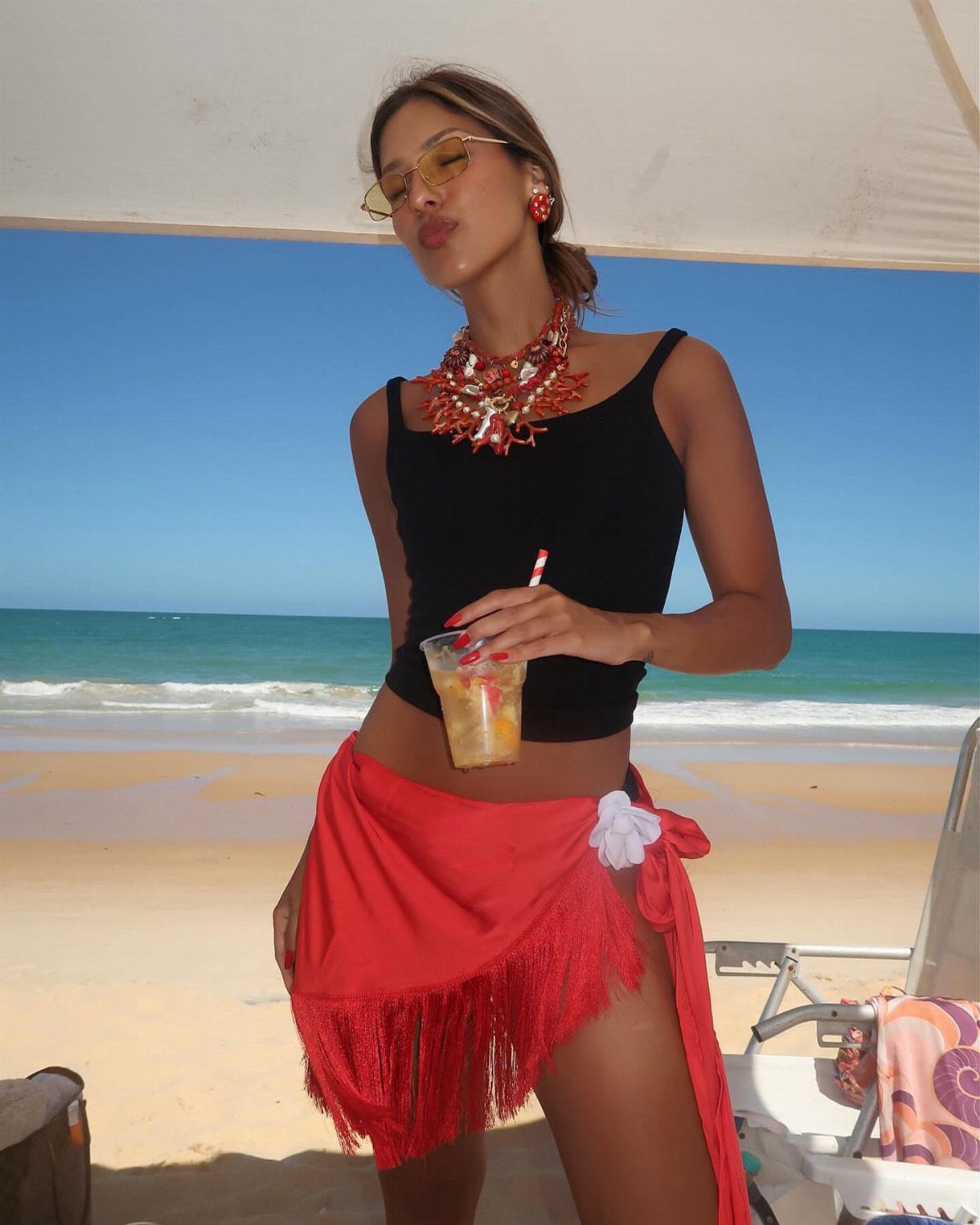 10 Chic Summer Outfits: Sandals, Linen, Dresses and More 7 Woman wearing a black tank top and a red fringe sarong on a sunny beach.