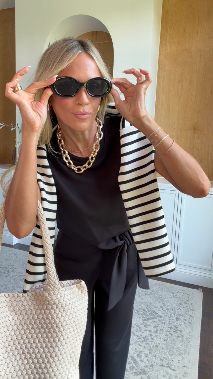 Chic Travel Outfits for Women: 14 Stylish Ideas for 2026 8 Woman wearing a black jumpsuit with a striped jacket and cream woven tote bag.