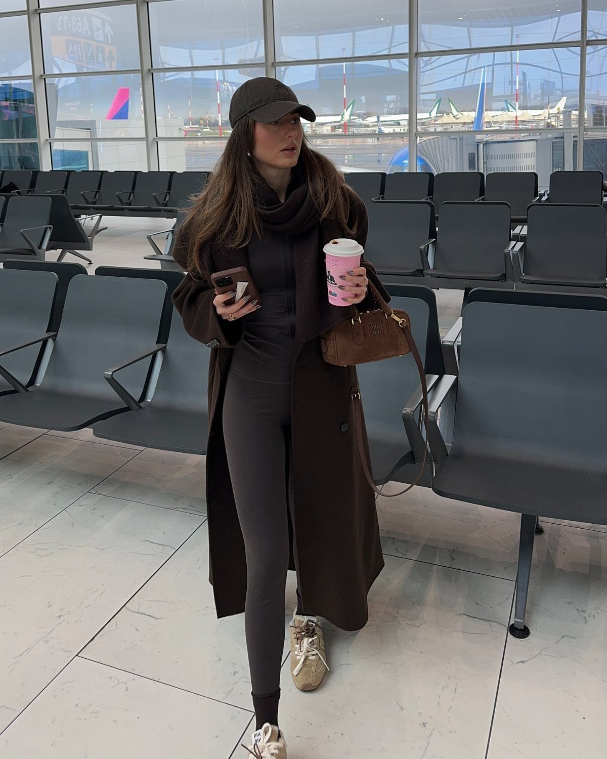 Chic Travel Outfits for Women: 14 Stylish Ideas for 2026 14 Woman in a long brown coat and black leggings standing in an airport terminal.
