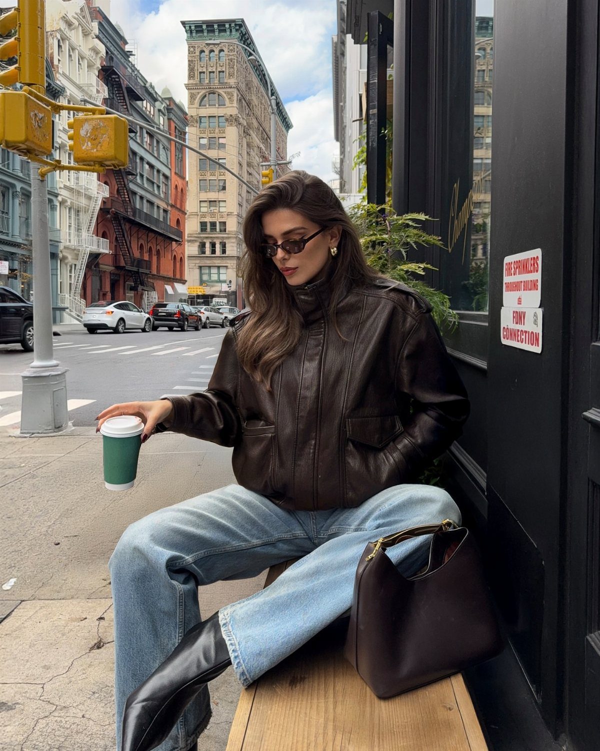 Chic Travel Outfits for Women: 14 Stylish Ideas for 2026 9 Woman in a brown leather jacket and wide-leg jeans sitting on a city bench.