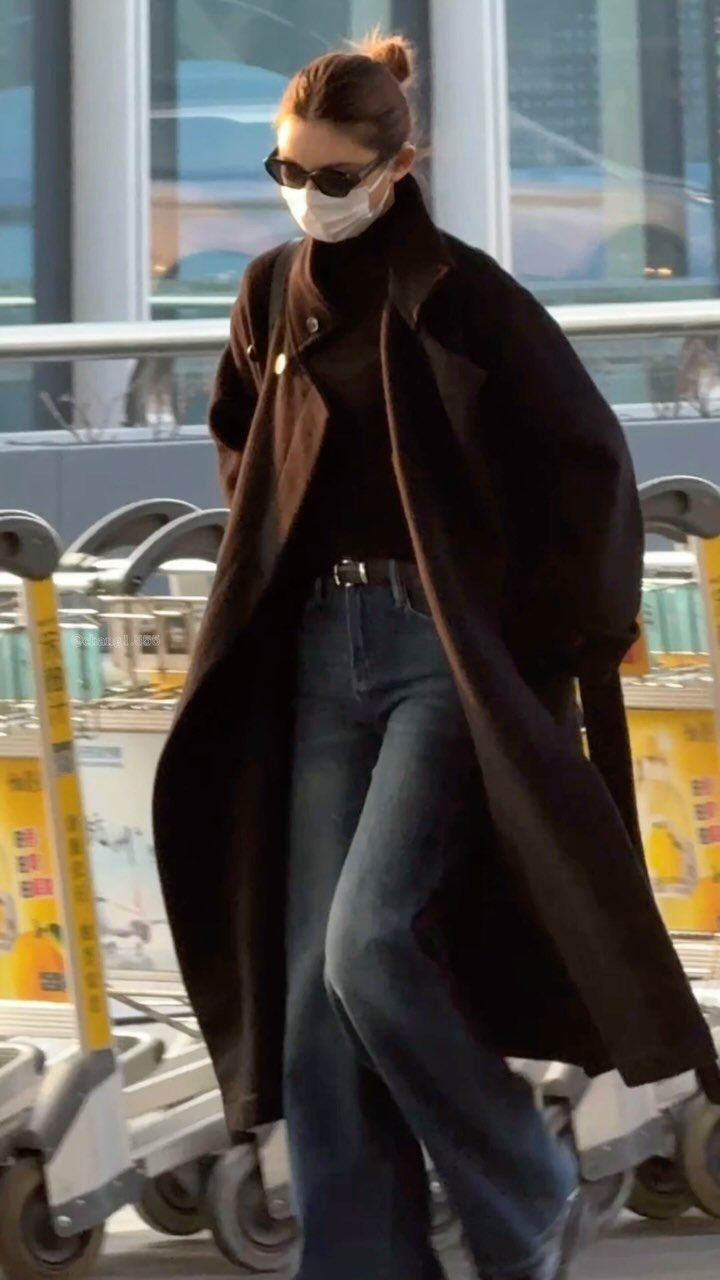 Chic Travel Outfits for Women: 14 Stylish Ideas for 2026 10 Woman in a long black coat and turtleneck walking through an airport terminal.