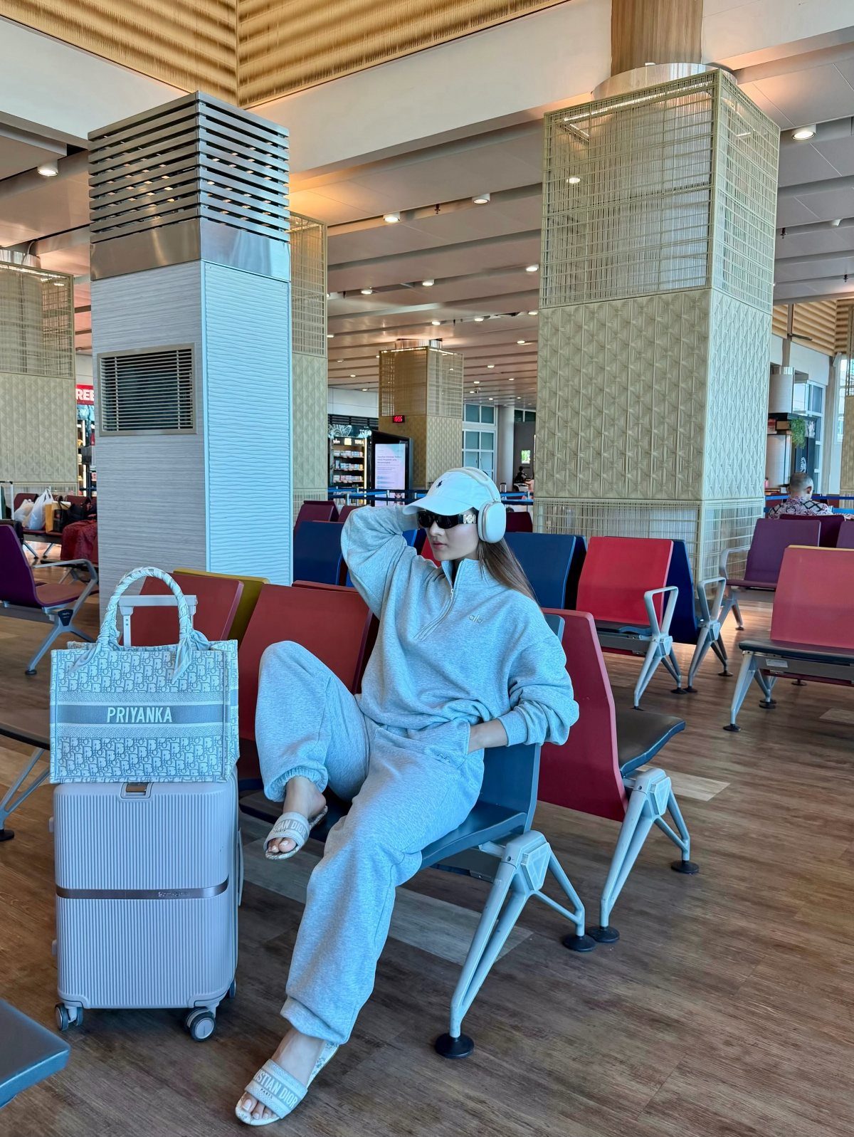 Chic Travel Outfits for Women: 14 Stylish Ideas for 2026 2 Woman in grey matching lounge set sitting in airport terminal with silver suitcase.