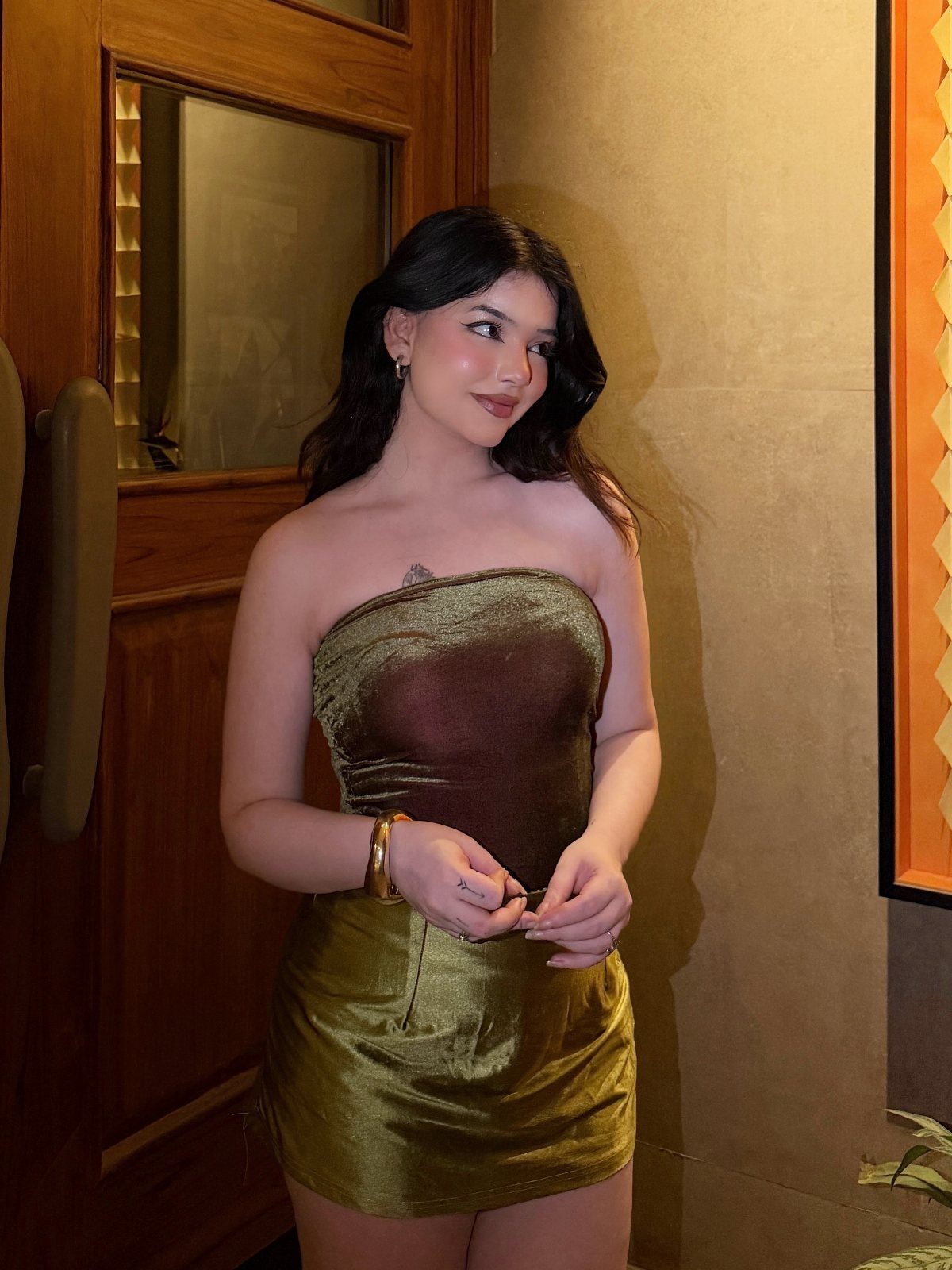 26 Classy Date Outfits From Casual to Elegant 24 Woman wearing a strapless metallic gold velvet mini dress standing by a wooden door.